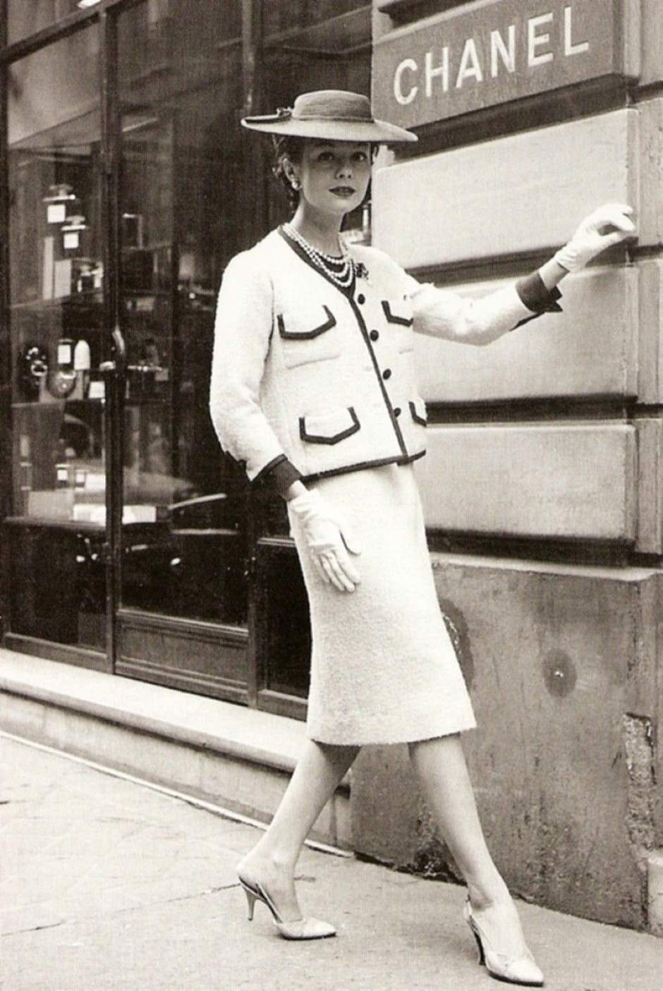 Foto em preto e branco de uma mulher bem vestida em frente a uma fachada da chanel